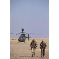 Portrait de deux militaires américains. En arrière-plan, un hélicoptère américain de reconnaissance et d'observation Bell OH-58 D Kiowa dans le désert en ZDO (zone de déploiement opérationnel) Olive.