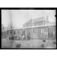 [Photographie de groupe rassemblant des enfants devant une école, accolée à une mairie, devant un bâtiment en reconstruction.]