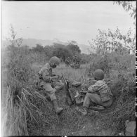 Des parachutistes vietnamiens se tiennent en position derrière un fusil-mitrailleur tandis que d'autres brancardent un blessé, au cours de l'opération Castor dans la vallée de Diên Biên Phu.