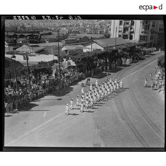 Plan en plongée du défilé d'une unité de la Marine nationale, sous le regard d'autorités militaires, civiles et de la population civile, à Alger.