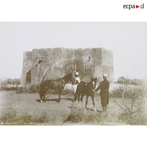 [Deux gardes africains et leurs chevaux devant un fort en terre crue.]