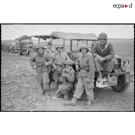 Vie quotidienne au 8e RIM. Portrait d'un groupe de soldats assis sur le devant d'une jeep. En arrière plan, un convoi militaire à l'arrêt. [légende d'origine]