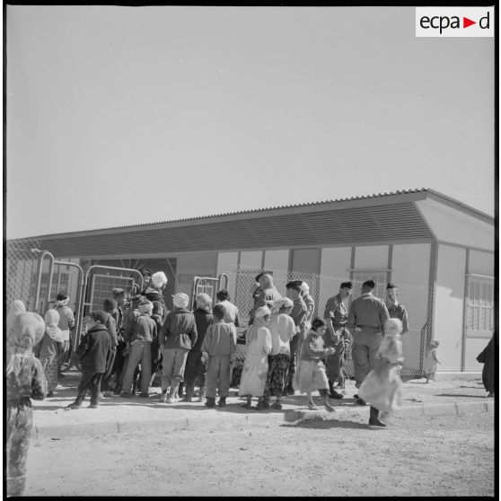 Devant un bâtiment, des soldats effectuent une distribution auprès des enfants de Wagram. [légende d'origine]