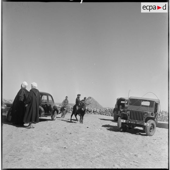 Dans un petit village, de jeunes garçons passent, à dos d'âne, entre des véhicules militaires à l'arrêt. [légende d'origine]