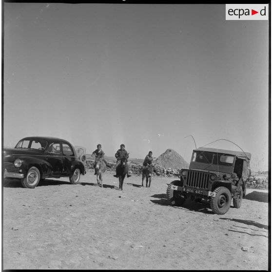 Dans un petit village, de jeunes garçons passent, à dos d'âne, entre des véhicules militaires à l'arrêt. [légende d'origine]