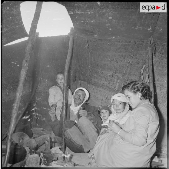 Portrait d'une famille à l'intérieur d'une khaïma. Sur la droite, une infirmière (EMSI) venue pour une séance d'auscultation boit le thé. [légende d'origine]