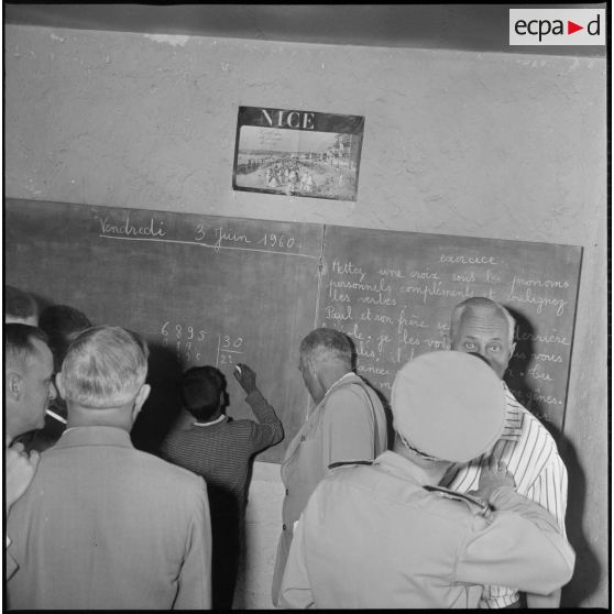 Les autorités civiles et militaires visitent une salle de classe. La date du vendredi 3 juin 1960 est inscrite sur le tableau de la classe. Au centre du tableau, un élève pose une division. [légende d'origine]