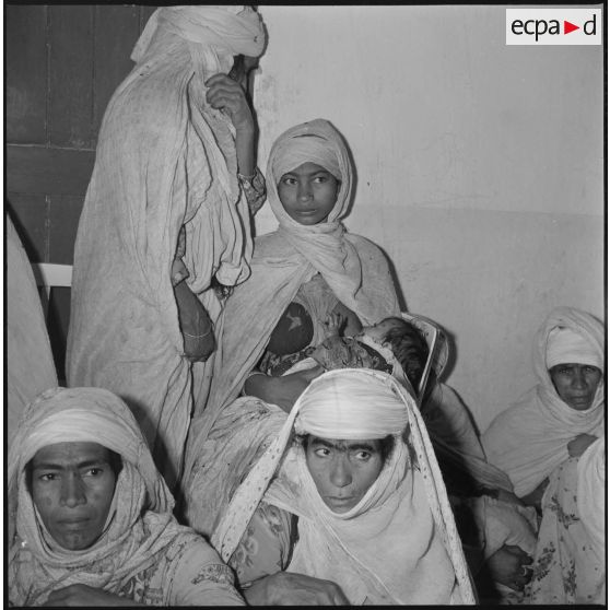 Portrait d'un groupe de femmes et d'enfants assis dans une salle d'attente. Un nourrisson tête au sein de sa mère. [légende d'origine]