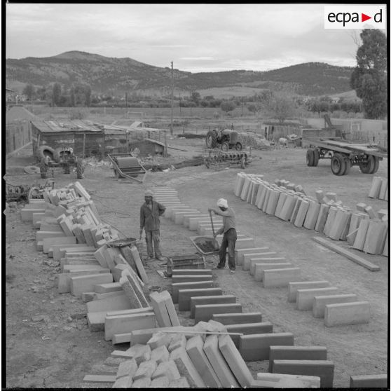 [Vue sur des matériaux de construction entreposés sur un terrain. Deux ouvriers sont en train de travailler pour la construction des nouvelles bâtisses du centre de regroupement de la ferme Cazorla.]