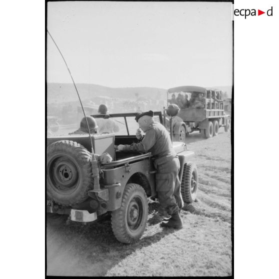 Un officier en liaison radio à bord d'une jeep. En arrière-plan, un convoi militaire. [légende d'origine]