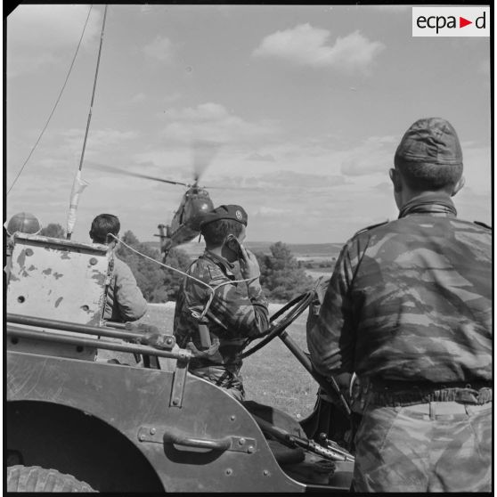 [Opération avec le commando Georges dans le secteur de Fenouane, près de la ferme Garrigues. Capitaine, accoudé au volant d'un véhicule de type jeep, en communication radio. Hélicoptère de type Sikorsky H-34 en vol.]