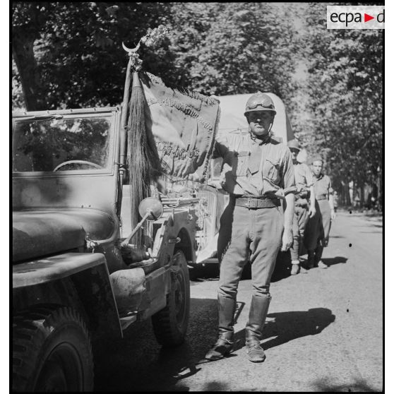 Portrait en pied d'un artilleur du 65e RAA (régiment d'artillerie d'Afrique) montrant un fanion de son régiment fixé à l'avant d'une jeep.