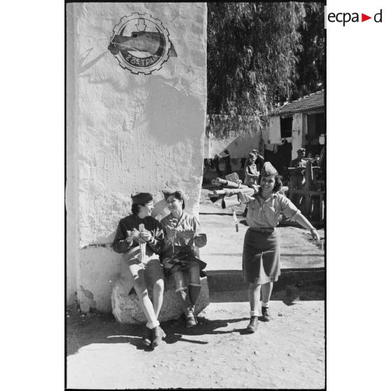 Assises sur un rocher, deux volontaires féminines conductrices à la 521e compagnie sanitaire tricotent pendant qu'une de leur camarade passe avec un brancard sur l'épaule.