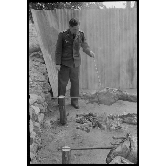 Un lieutenant d'un service administratif de la Luftwaffe surveille la cuisson de la viande lors d'un méchoui organisé au sein d'une unité de reconnaissance de l'armée de l'Air allemande (Aufklärungsgruppe).