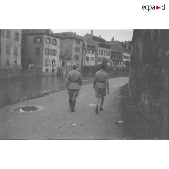 [Années 1930. Deux militaires photographiés de dos marchant au bord d'une rivière dans une ville].