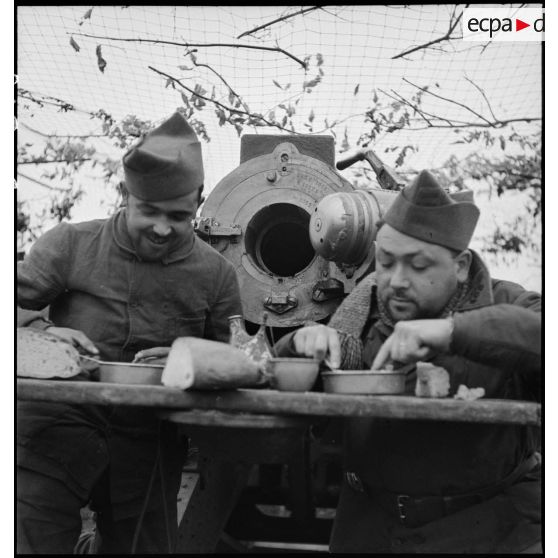 Deux artilleurs du 185e RALT (régiment d'artillerie lourde tractée) se restaurent près de la pièce qu'ils servent, un canon de 145/155 mm m1916, culasse ouverte.