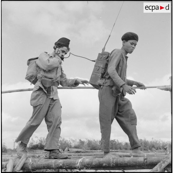 Le transmetteur radio du bataillon de marche indochinois (BMI) et son porteur vietnamien sur un pont de bambous au cours de l'opération Marécages.