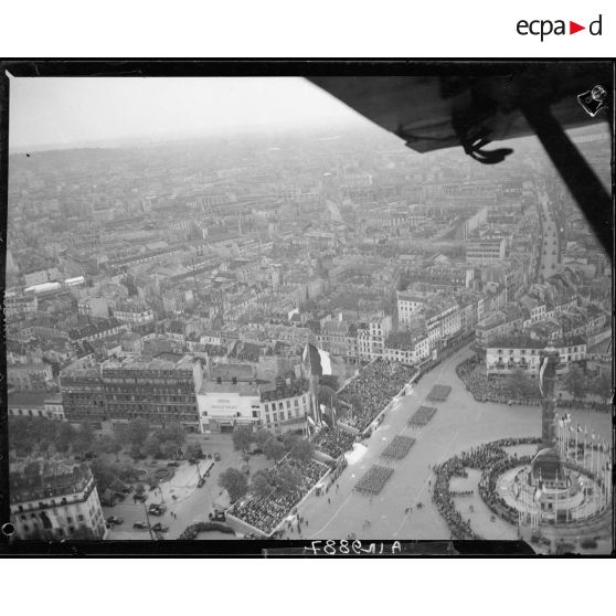 Vue aérienne des cérémonies du 14 juillet 1946 à Paris. Des troupes à pied défilent sur la place de la Bastille devant les tribunes. En arrière-plan, le cinéma Bastille Palace.