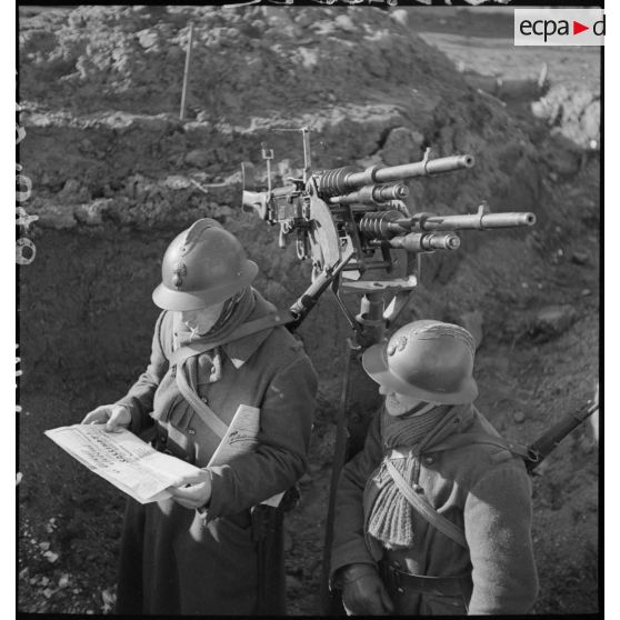 Sur le terrain, deux fantassins, servants de deux mitrailleuses jumelées Hotchkiss modèle 1914 de 8 mm, sur affût antiaérien, lisent un numéro du journal de campagne intitulé Le tireur debout.