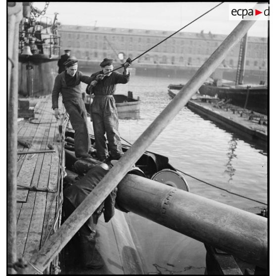 Chargement, à l'aide d'un palan, de torpilles dans les tubes lance-torpilles d'une tourelle orientable d'un sous-marin, à quai dans un port.