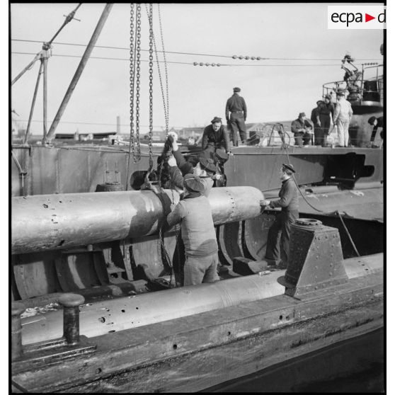 Chargement, à l'aide d'un palan, de torpilles dans les tubes lance-torpilles d'une tourelle orientable d'un sous-marin, à quai dans un port.