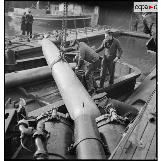 Chargement, à l'aide d'un palan, de torpilles dans les tubes lance-torpilles d'une tourelle orientable d'un sous-marin, à quai dans un port.