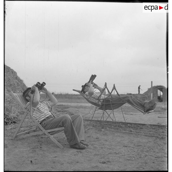 Des marins, appartenant à la 4e batterie de 90 mm contre avions, batterie mobile de défense antiaérienne de la Marine, observent le ciel à l'aide de jumelles, installés dans des chaises longues ou des sièges suspendus.