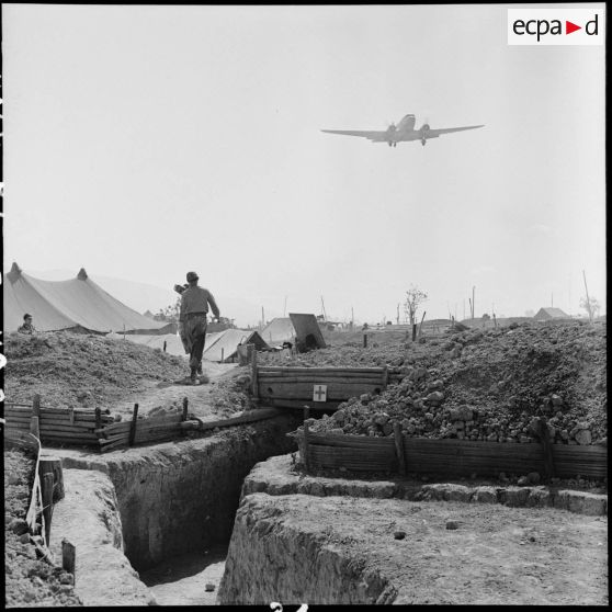 Un avion de transport Dakota survole le camp retranché de Diên Biên Phu dont on aperçoit l'entrée d'une antenne chirurgicale.