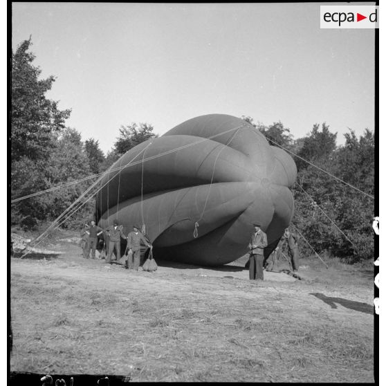 Des aérostiers maintiennent au sol un ballon de protection photographié en plan général de trois quarts avant.