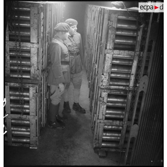 Deux officiers, dont un lieutenant, inspectent des obus de 75 mm stockés sur chariot métallique dans l'une des casemates à munitions de l'ouvrage de Velosne.