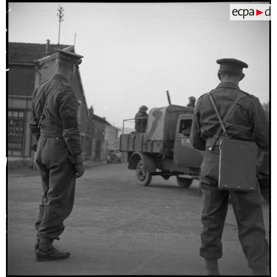 Deux soldats de la BEF, photographiés de dos dans un village, regardent passer un convoi.