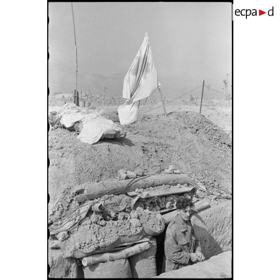 Soldat dans une tranchée à l'entrée d'une antenne chirurgicale du camp retranché de Diên Biên Phu, sur laquelle flotte le drapeau blanc à Croix-rouge.