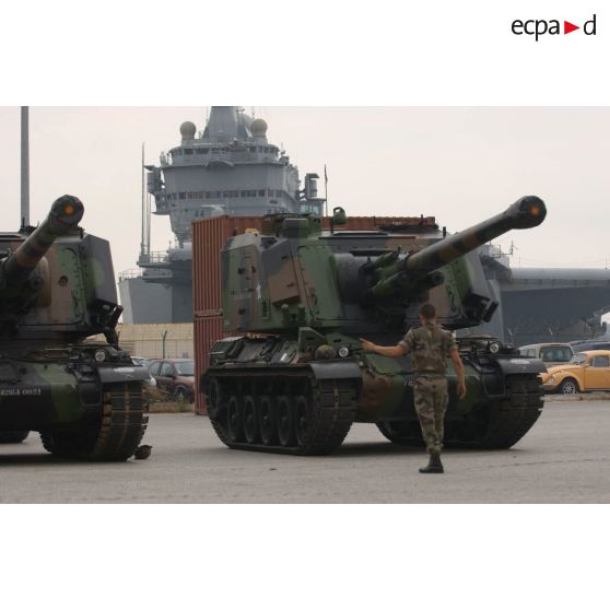 Des AU-F1 (canon automoteur de 155 mm à grande cadence de tir modèle F1) du 1er RAMa (régiment d'artillerie de marine) stationnés sur un parking de l'arsenal de Toulon avant l'embarquement pour le Liban, avec en arrière-plan le porte-avions nucléaire Charles-de-Gaulle à quai.