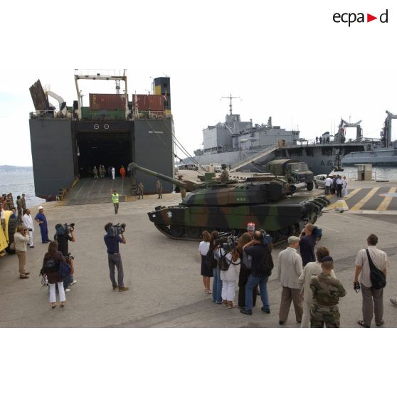 Le char Leclerc Bayeux du 6/12e RC (régiment de cuirassiers), à l'embarquement pour le Liban, monte la rampe d'accès du cargo affrété Fast Arrow sur le port de Toulon, guidé par un personnel du 601e RCR (régiment de circulation routière), en présence de caméramen et de photographes. En arrière-plan, le BAP (bâtiment-atelier polyvalent) de la Marine nationale Jules Verne à quai.