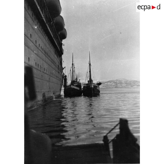 Deux bateaux de pêche accostent le flanc d'un paquebot /transport de troupes dans un fjord.