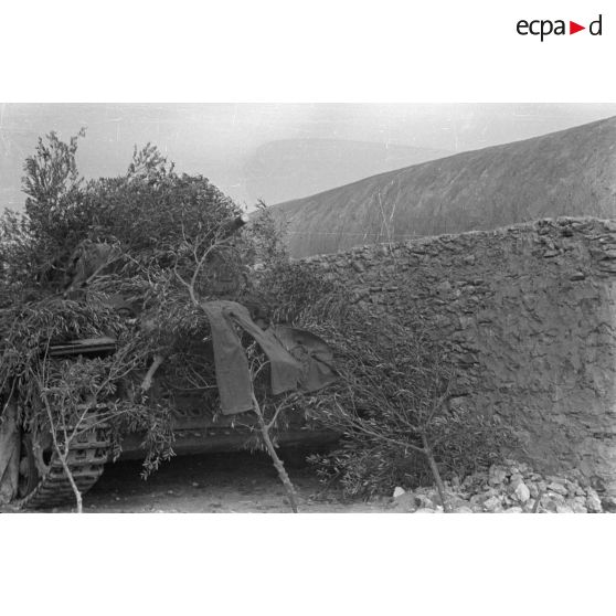 Un char Panzer III (Pz-III Ausf-L) est camouflé sous des branchages près d'une habitation de pierre.