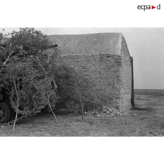 Un char Panzer III (Pz-III Ausf-L) est camouflé sous des branchages près d'une habitation de pierre.