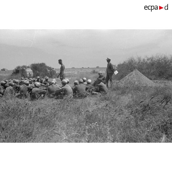 Des soldats assis à même la terre, en demi-cercle, écoutent un cadre torse nu. Tentes et véhicules divers.