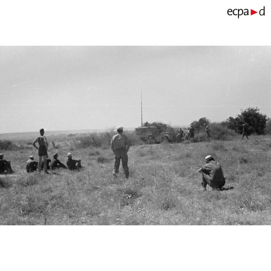 Des soldats assis à même la terre, en demi-cercle, écoutent un cadre torse nu. Tentes et véhicules divers.