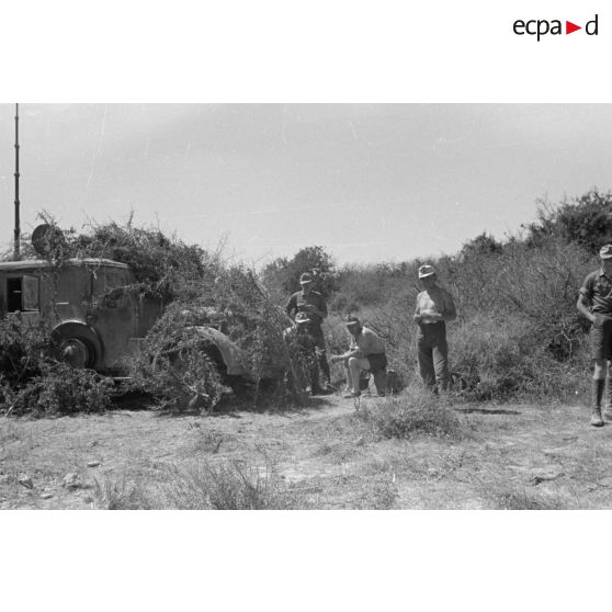 Des soldats assis à même la terre, en demi-cercle, écoutent un cadre torse nu. Tentes et véhicules divers.