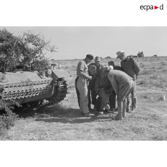 Un groupe de soldats du Panzer Regiment 5 (Pz.Rgt-5) entoure un camarade qui écrit ou lit un texte près d'un char Panzer III (Pz-III Ausf J) camouflé par de la végétation.