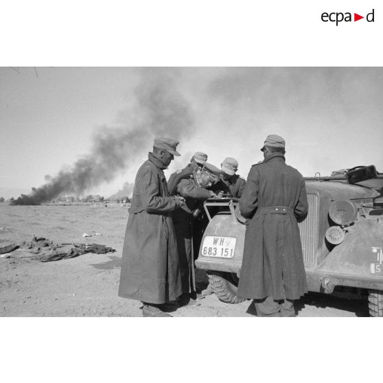 A l'appui du capot d'une Horch immatriculée WH 693 151, le général (General der Panzertruppe) Erwin Rommel, accompagné du Generalmajor Gause, donne des ordres au milieu de véhicules en flamme.