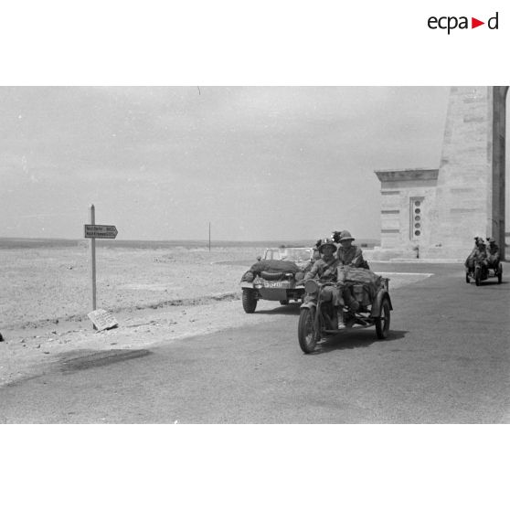 Une voiture allemande Kübelwagen et des mototricycles italiens d'une unité de Bersaglieri passent sous l'Arco dei Fileni sur la via Balbia.