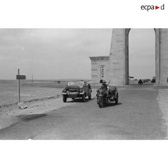 Voiture Kübelwagen et mototricycle italien d'une unité de Bersaglieri passant sous l'Arco dei Fileni sur la via Balbia.