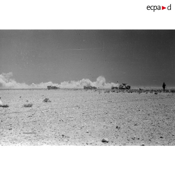 La colonne de véhicules face à un nuage de poussière soulevé par ce qui semble être les explosions d'un tir d'artillerie ou par un vent de sable.