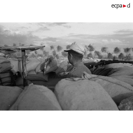 Un soldat observe le terrain depuis une tranchée,. des fusils Mauser (KAR-98k) sont posés sur les sacs de sable du parapet.