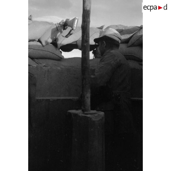 Un soldat observe le terrain depuis une tranchée,. des fusils Mauser (KAR-98k) sont posés sur les sacs de sable du parapet.