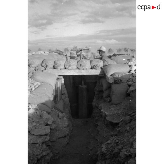 Un soldat observe le terrain depuis une tranchée,. des fusils Mauser (KAR-98k) sont posés sur les sacs de sable du parapet.