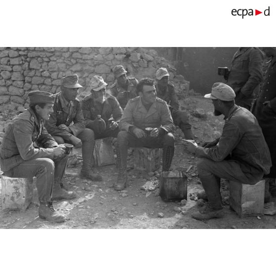 Un groupe de soldats parle et se restaure, près d'un mur en partie éboulé.