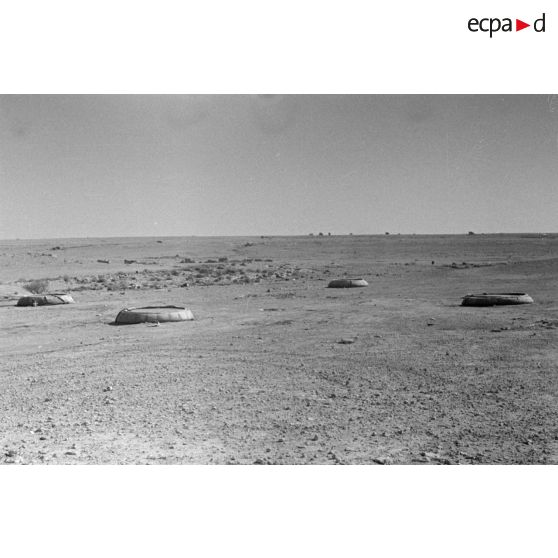 Vue du désert et de quatre bacs souples, au loin passent des véhicules.